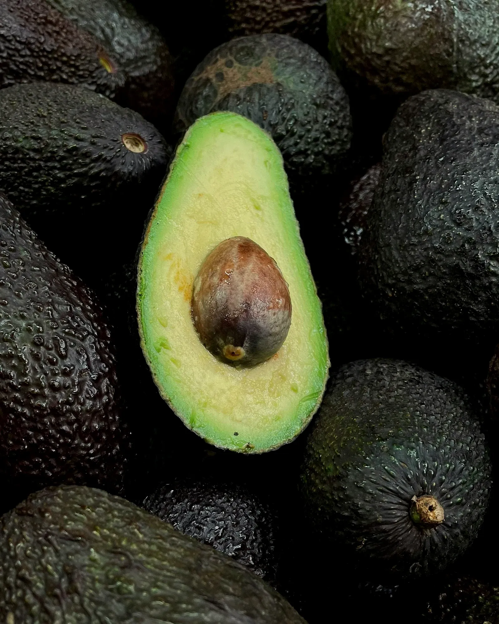 A halved avocado with its stone, lying among whole dark-skinned Hass avocados.